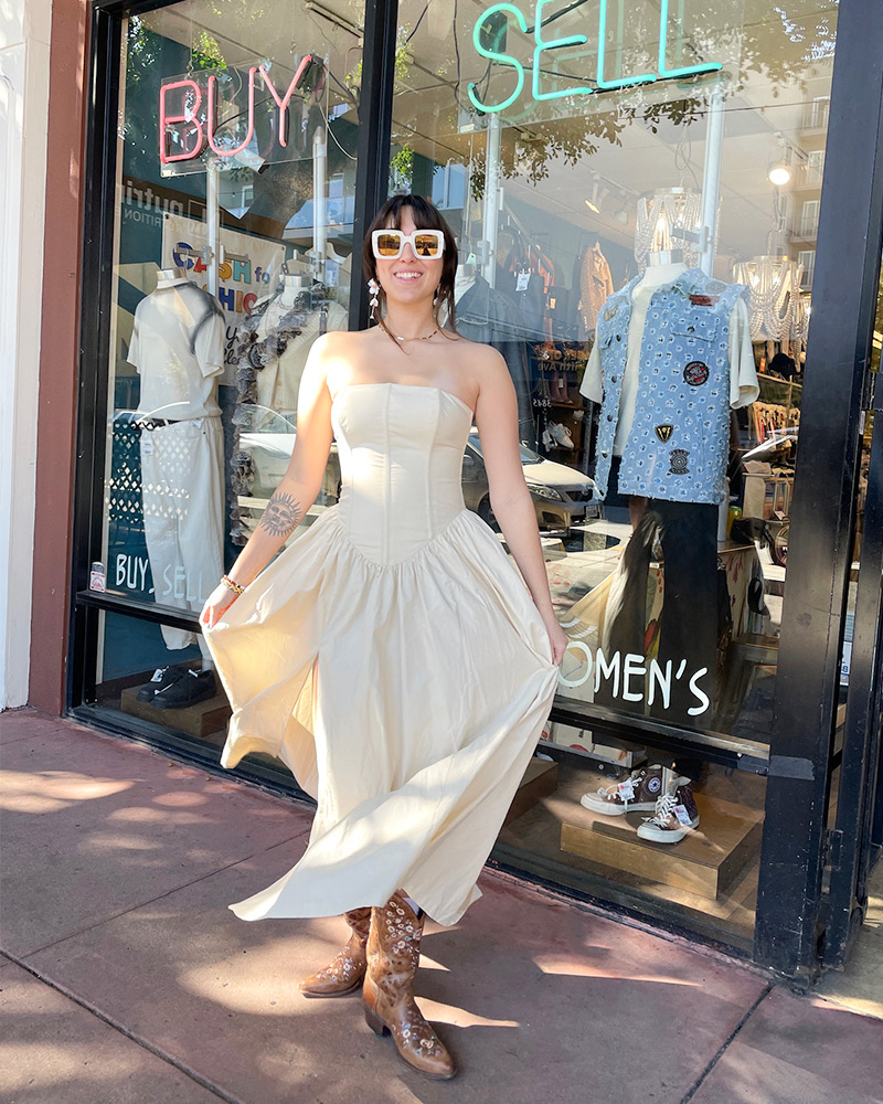 Woman wearing cream strapless dress and brown western boots stands in front of shop window